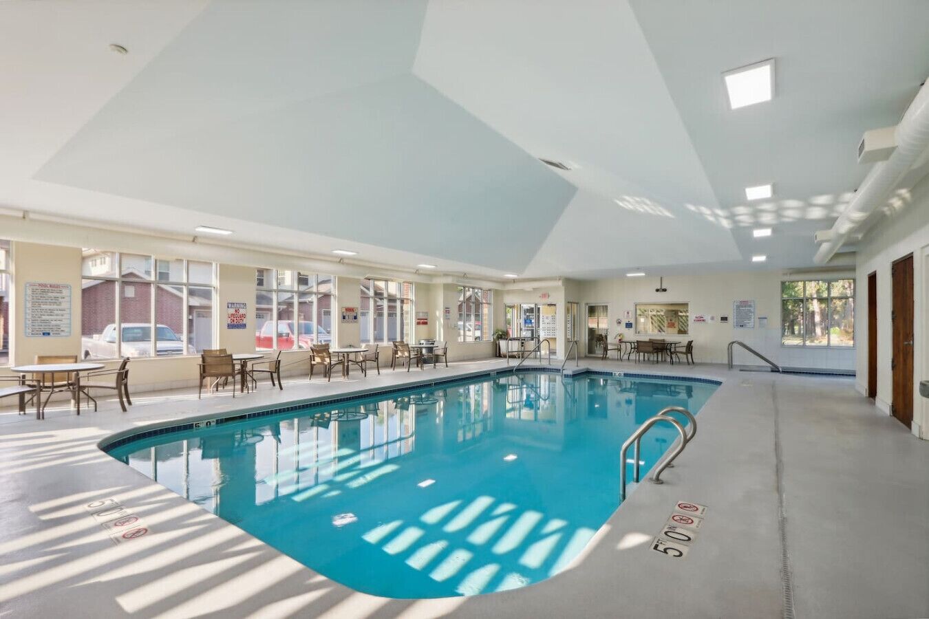 An indoor swimming pool with clear water, surrounded by tables and chairs, and large windows allowing natural light.