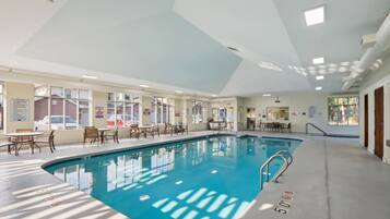An indoor swimming pool with clear water, surrounded by tables and chairs, and large windows allowing natural light.