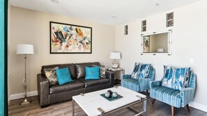 A living room with a dark leather sofa, two patterned armchairs, a white coffee table, and a large abstract painting on the wall.