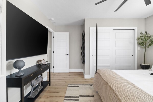 A modern bedroom with a large bed, a TV mounted on the wall, a console table with decorative items, and a potted plant.