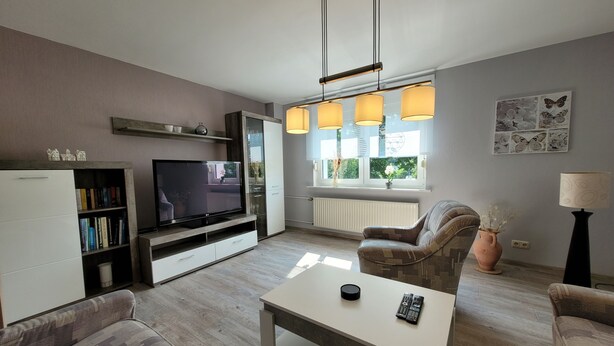 Una sala de estar moderna con un televisor de pantalla plana, una mesa de centro blanca, un sofá estampado, una estantería y una ventana con luz natural.