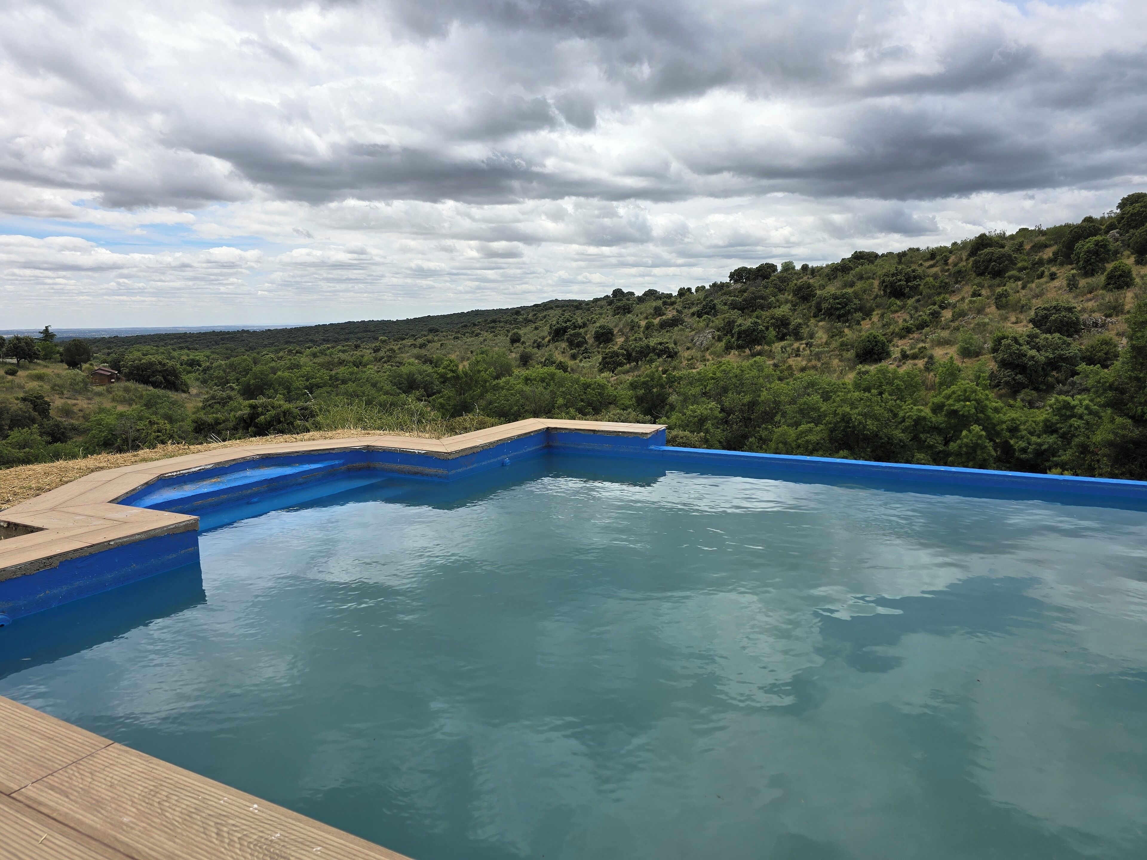 A swimming pool with a wooden deck, surrounded by lush greenery and hills.