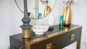 A dark wooden dresser with brass handles, a white bowl, a teal vase, and a mirror reflecting flowers.