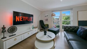 A modern living room with a large flat-screen TV displaying 'NETFLIX', a white console table, a round coffee table, a black leather sofa, and a large window with a view of the outside.
