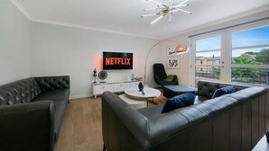 A modern living room with a leather sofa, a coffee table, a TV displaying 'NETFLIX', and a large window offering a view of the city.