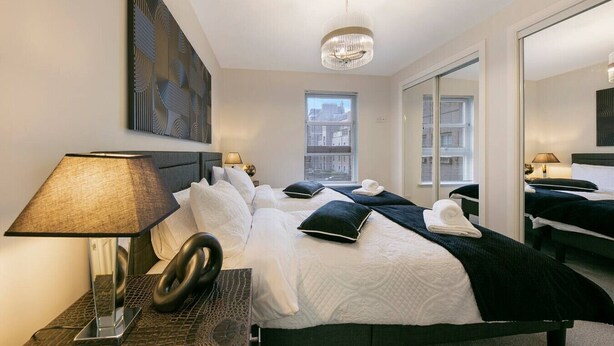 A modern hotel room with a large bed, a bedside lamp, a mirror, and a city view.