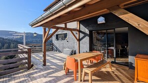 Une terrasse en bois avec une table et des chaises, une vue sur les montagnes et un bâtiment à la façade sombre, doté d’une fenêtre.