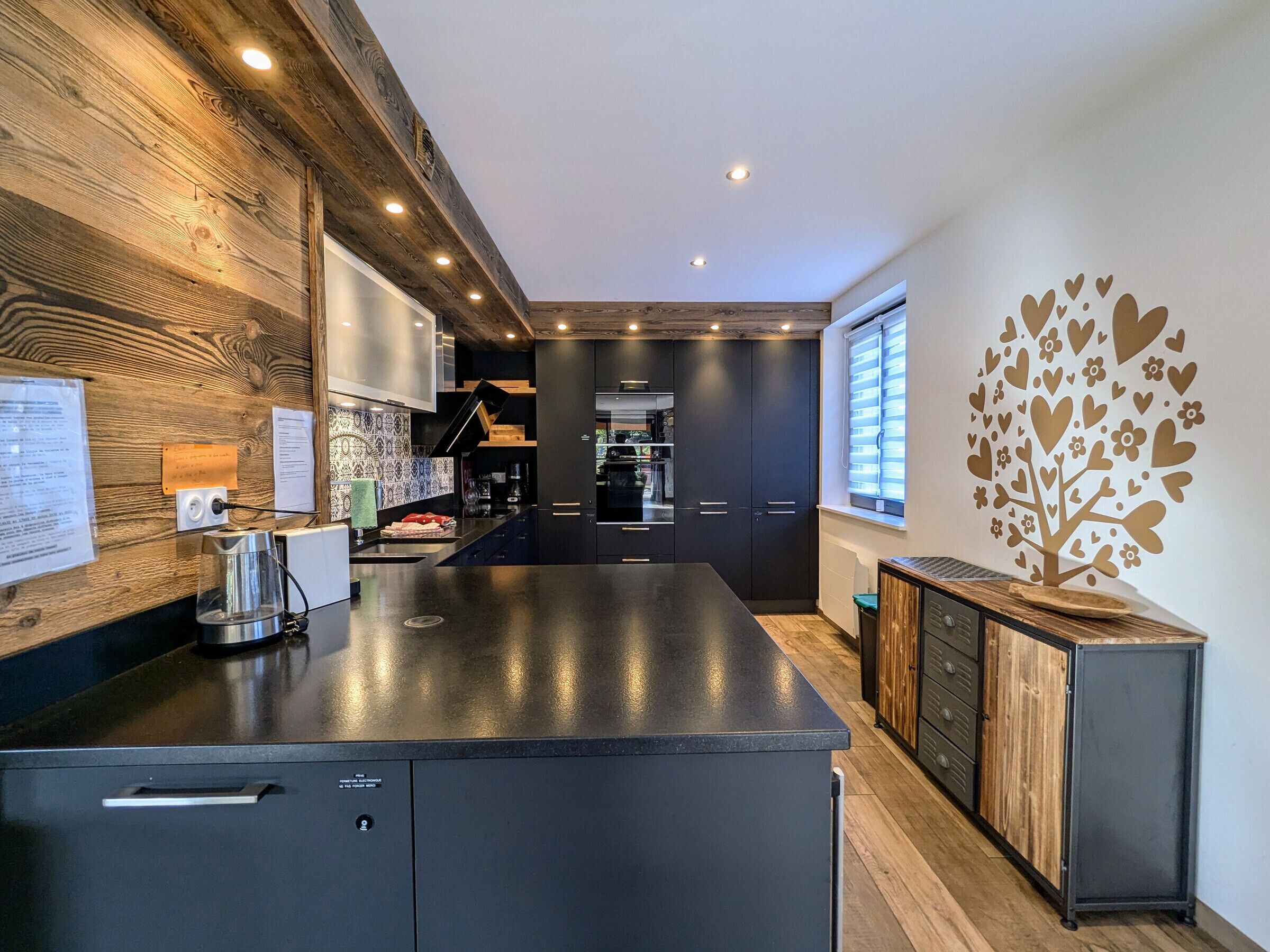 A modern kitchen with a central island, dark cabinetry, and a decorative tree wall art.