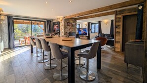 Un vaste espace dédié à la salle à manger, avec une longue table et des chaises modernes, un mur décoratif en pierre et un téléviseur en arrière-plan.