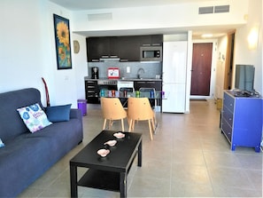 A modern living room with a sofa, a coffee table, and two chairs. A kitchen with dark cabinets, a microwave, and a refrigerator is visible in the background.