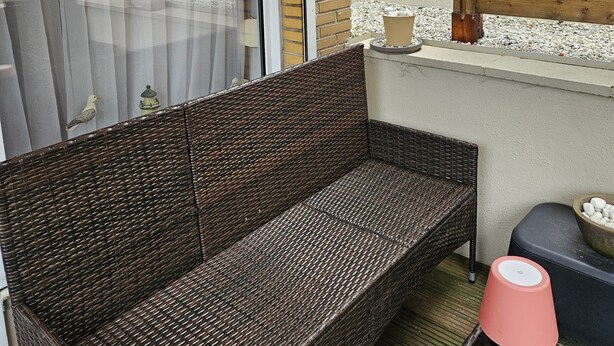 A wicker sofa with a cushion, a small table with a lamp, and a window with a curtain. - Quiet and relaxing apartment in Coxyde, 5 minutes walk from the beach (Koksijde)