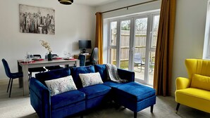 A living room with a blue sectional sofa, a yellow armchair, a dining area with a table and chairs, and a large window with curtains.