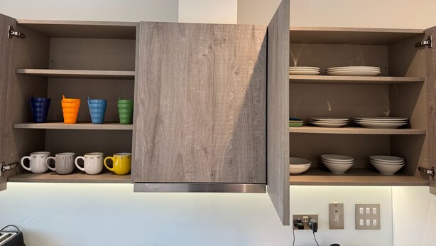A modern kitchen with open wooden cabinets, shelves with colorful mugs, and white plates.