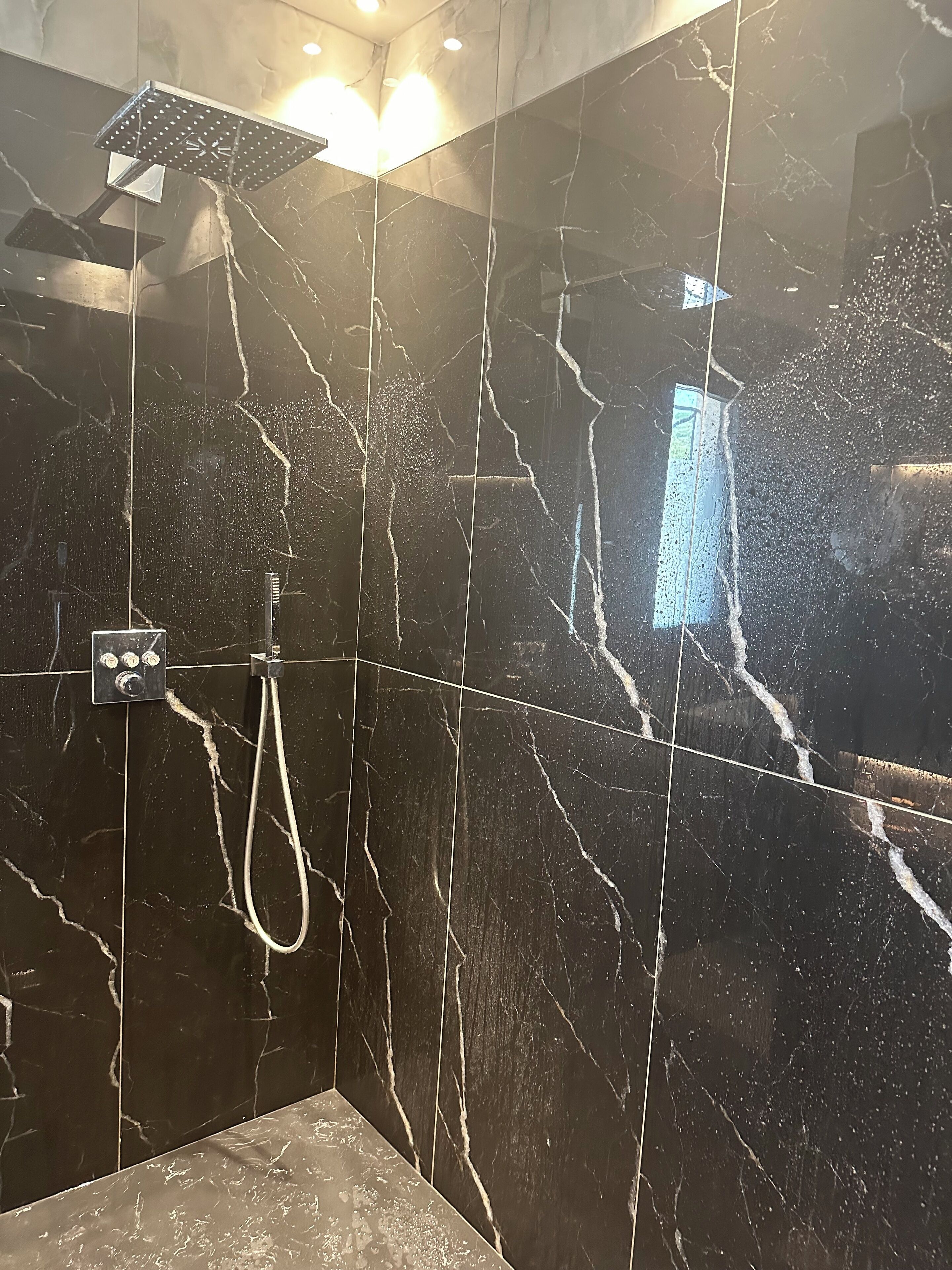 A modern bathroom with black marble tiles, a showerhead, and a control panel.