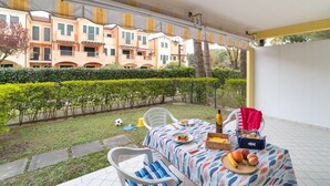 An outdoor patio with a table set for a meal, surrounded by a neatly trimmed hedge and a row of buildings in the background. - Beautiful flat with large garden and terrace (Bibione)