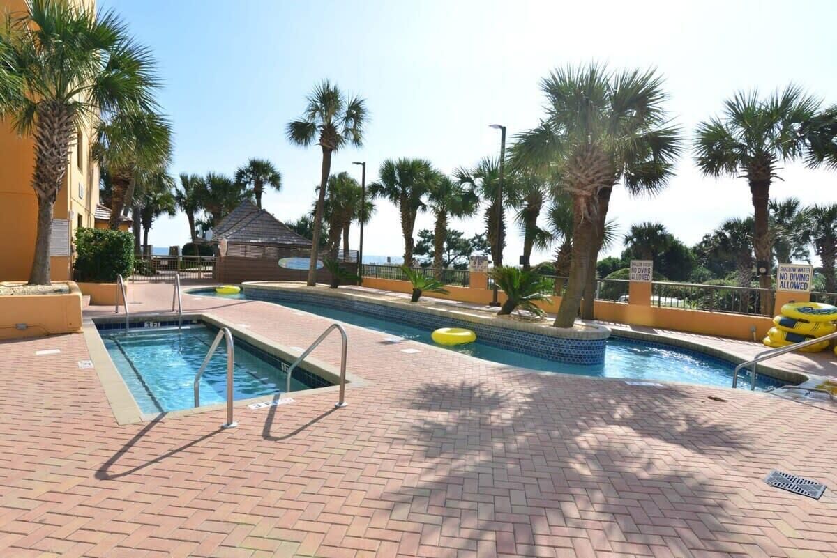 A swimming pool with a brick deck, palm trees, and a small building in the background.
