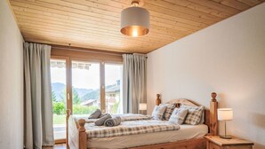 Ein hölzernes Bett mit Blick auf Berge, ein kleiner Nachttisch mit Lampe und ein großes Fenster mit Vorhängen.