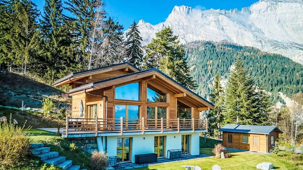 Eine moderne Holzhütte mit Balkon, umgeben von einem gepflegten Garten und einer Berglandschaft.