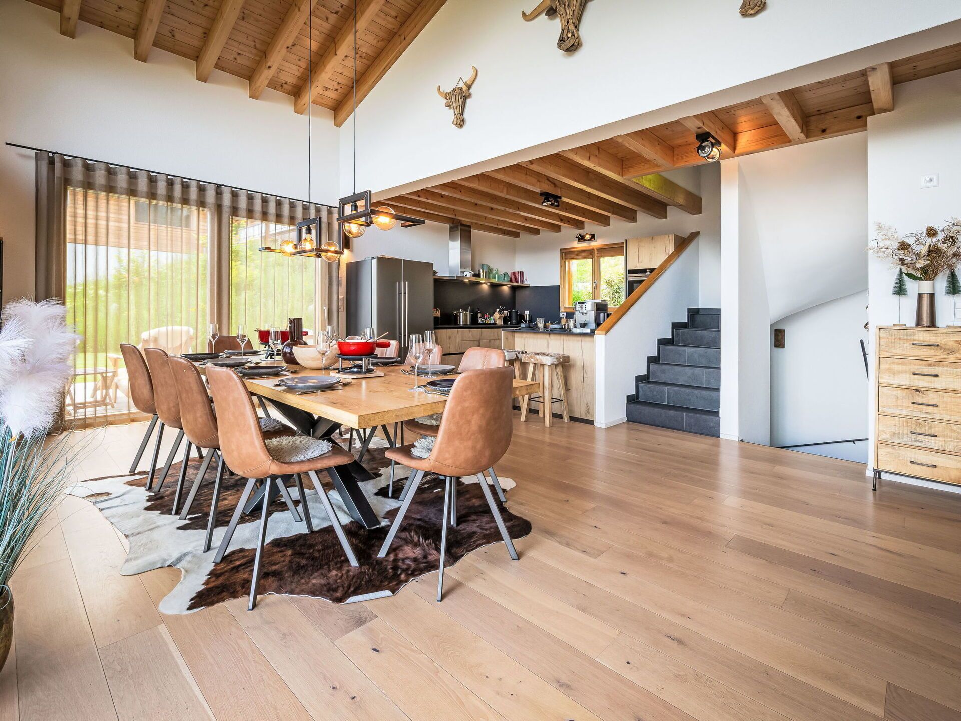A spacious dining area with a wooden table and chairs, a kitchen in the background, and a staircase leading to an upper level.