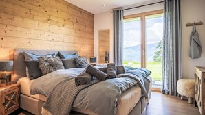 Ein Schlafzimmer mit Holzwand, einem großen Bett mit vielen Kissen, einem Nachttisch und Blick ins Freie durch eine Schiebetür.