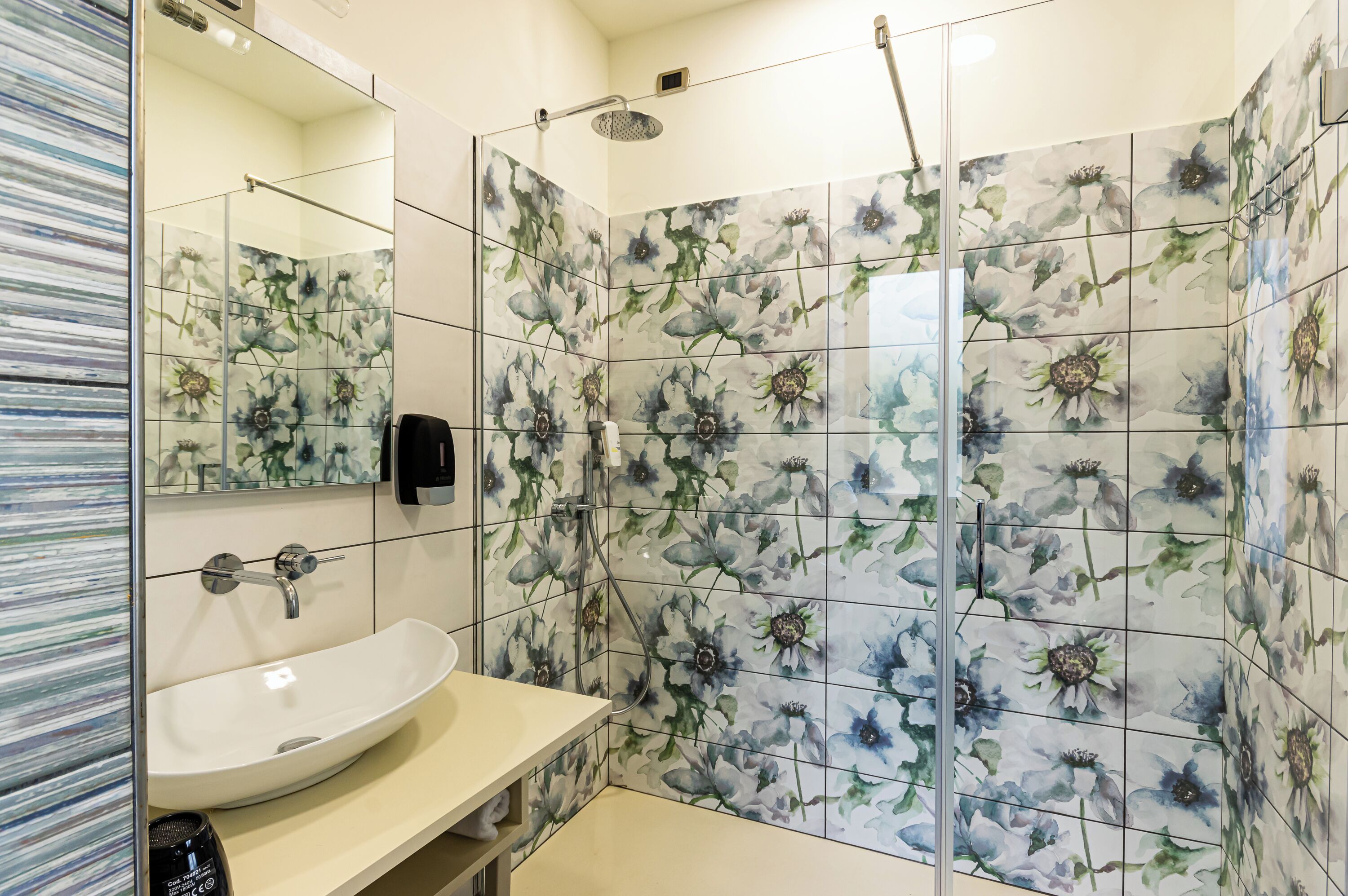 Ein Badezimmer mit einem weißen Waschbecken, einer Duschkabine aus Glas und Fliesen mit floralem Muster.