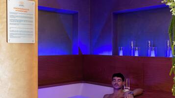 Um homem relaxando em uma banheira de madeira com um copo de vinho, cercado por iluminação azul e uma placa na parede.