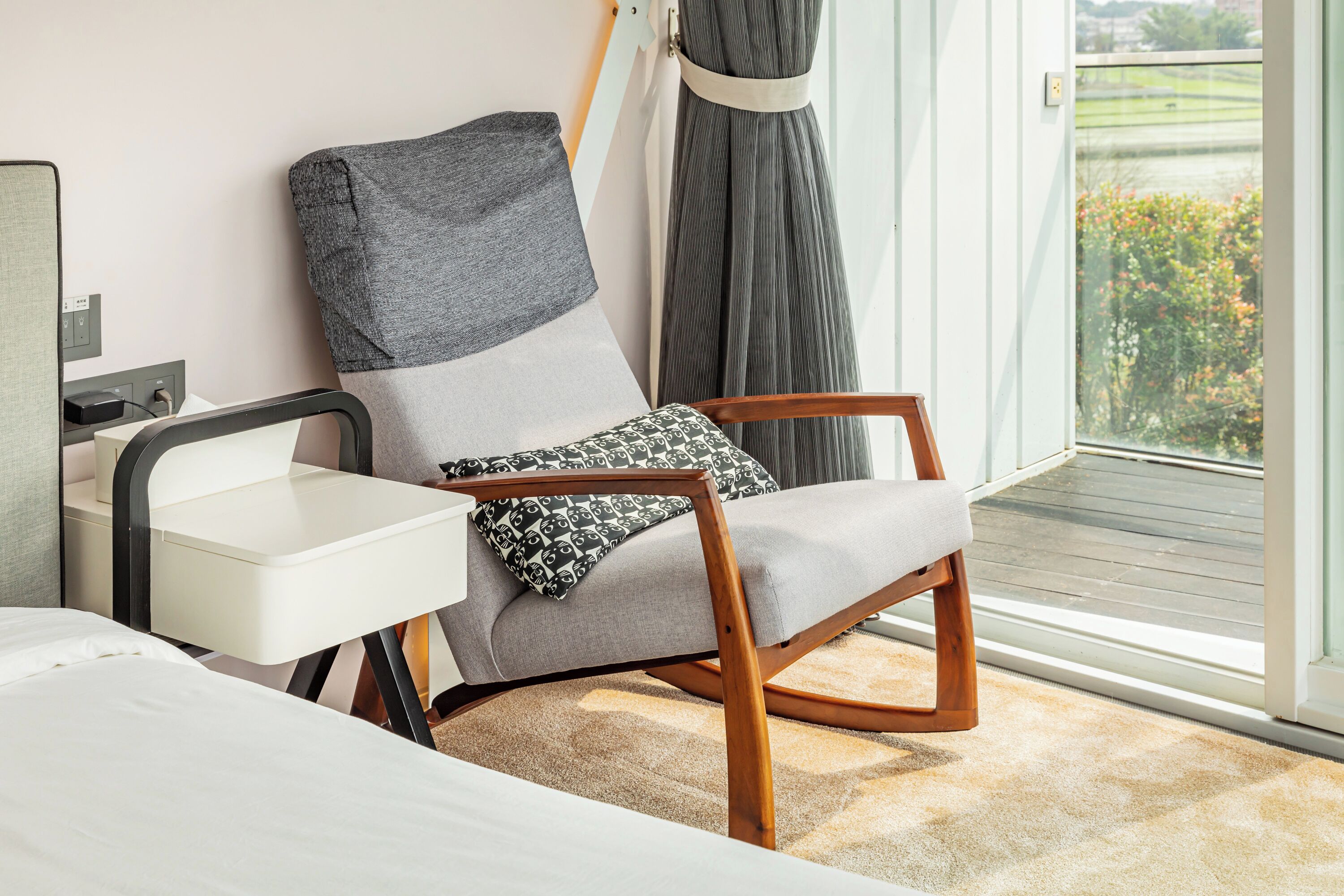 Una habitación moderna con un sofá gris, una silla de madera, una mesita blanca y un amplio ventanal con vistas a zonas verdes.