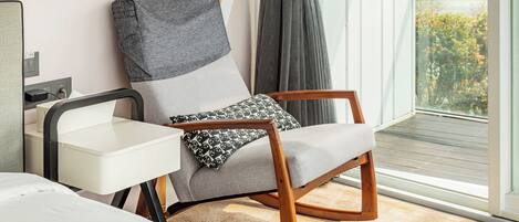 Una habitación moderna con un sofá gris, una silla de madera, una mesita blanca y un amplio ventanal con vistas a zonas verdes.