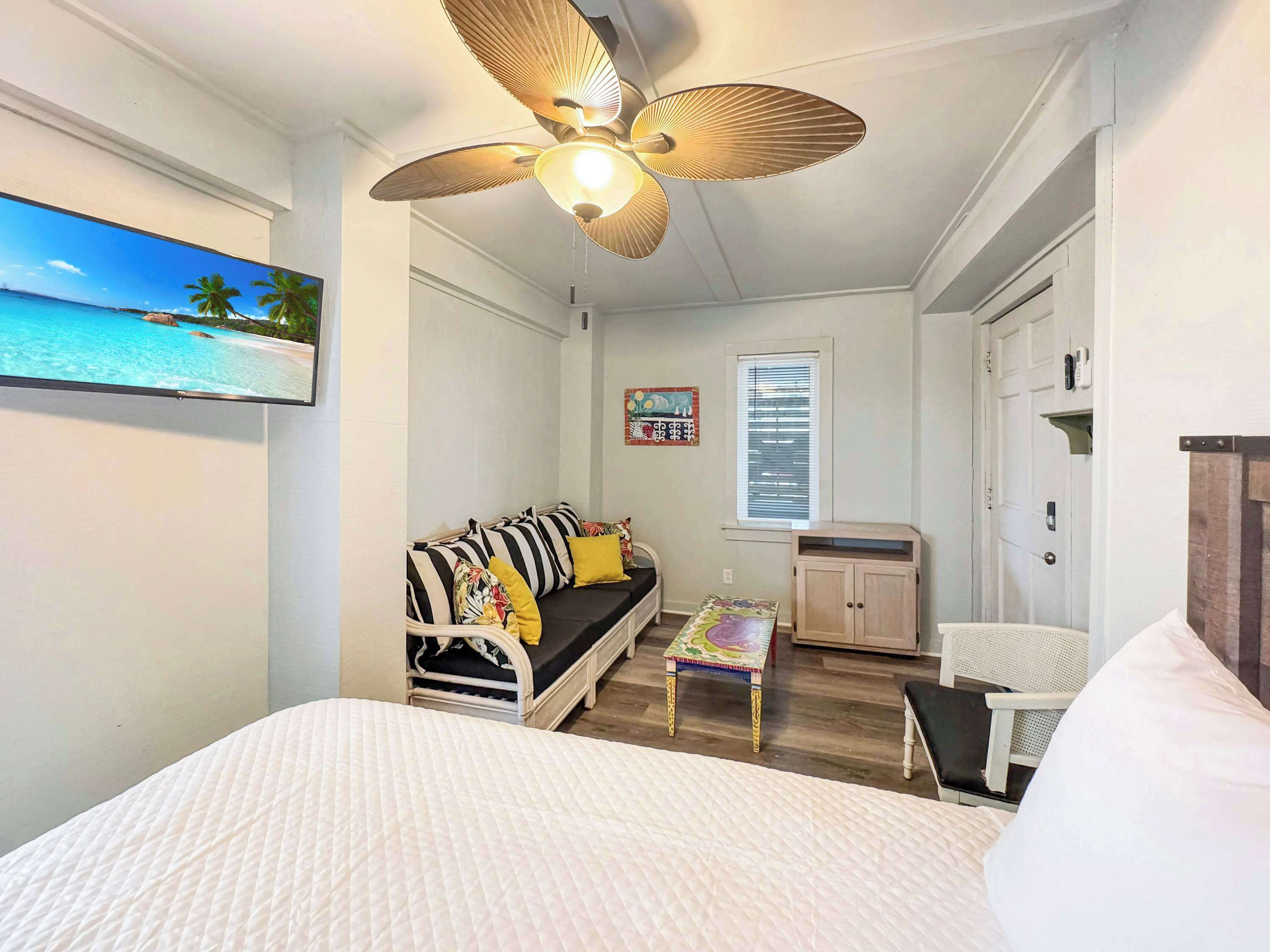 A bedroom with a bed, a sofa, a small table, a ceiling fan, a window with blinds, and a door.