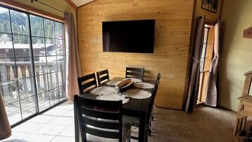 Un coin repas avec un mur en bois, un téléviseur à écran plat fixé au-dessus d’une table entourée de quatre chaises, et une grande fenêtre donnant sur des arbres et des immeubles.