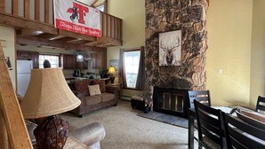 Ein geräumiges Wohnzimmer mit Kamin aus Stein, einer hohen Decke mit freiliegenden Balken, einer Küche im Hintergrund und einem Banner für Texas Tech Red Raiders.