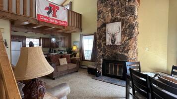 Amplia sala de estar con chimenea de piedra, techo alto con vigas a la vista, cocina al fondo y un banderín de Texas Tech Red Raiders.