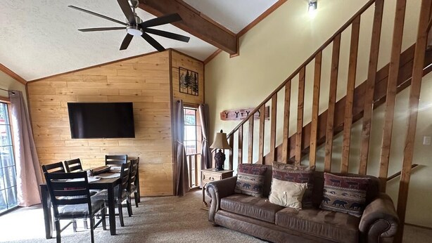 Ein Wohnzimmer mit Holzbalkendecke, einem flachbildfernseher an der Wand, einem braunen Sofa mit gemusterten Kissen, einem Esstisch mit Stühlen und einer Treppe mit Holzbalken.
