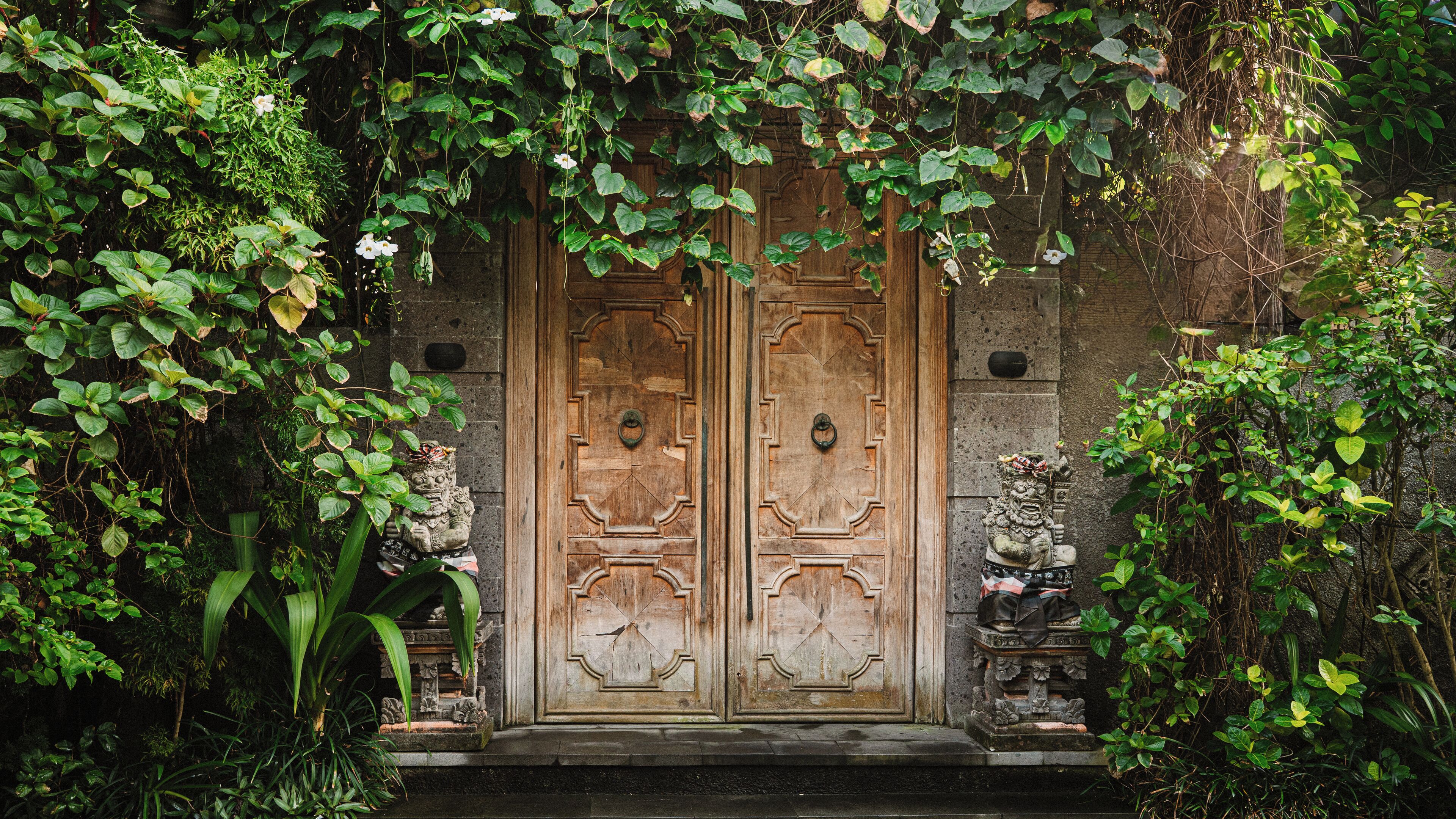 Une porte en bois ornée de sculptures complexes, flanquée de statues de pierre et entourée d’une végétation luxuriante.