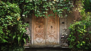 Una porta in legno finemente intagliata, affiancata da statue in pietra e circondata da rigogliosa vegetazione.