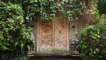Une porte en bois ornée de sculptures complexes, flanquée de statues de pierre et entourée d’une végétation luxuriante.