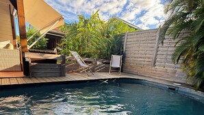 En swimmingpool med træterrasse og liggestole, omgivet af et træhegn og tropiske planter.