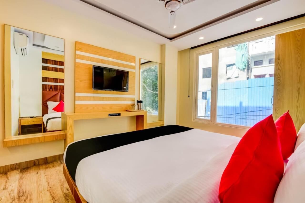 A modern hotel room with a large bed, a TV mounted on a wooden panel wall, a small desk with a chair, and a sliding glass door leading to a balcony.