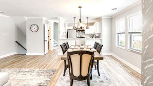 A spacious dining area with a wooden table and chairs, a kitchen in the background, and a clock on the wall.