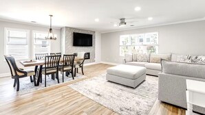 A spacious living room with a dining area, a grey sofa, and a flat-screen TV mounted on the wall.