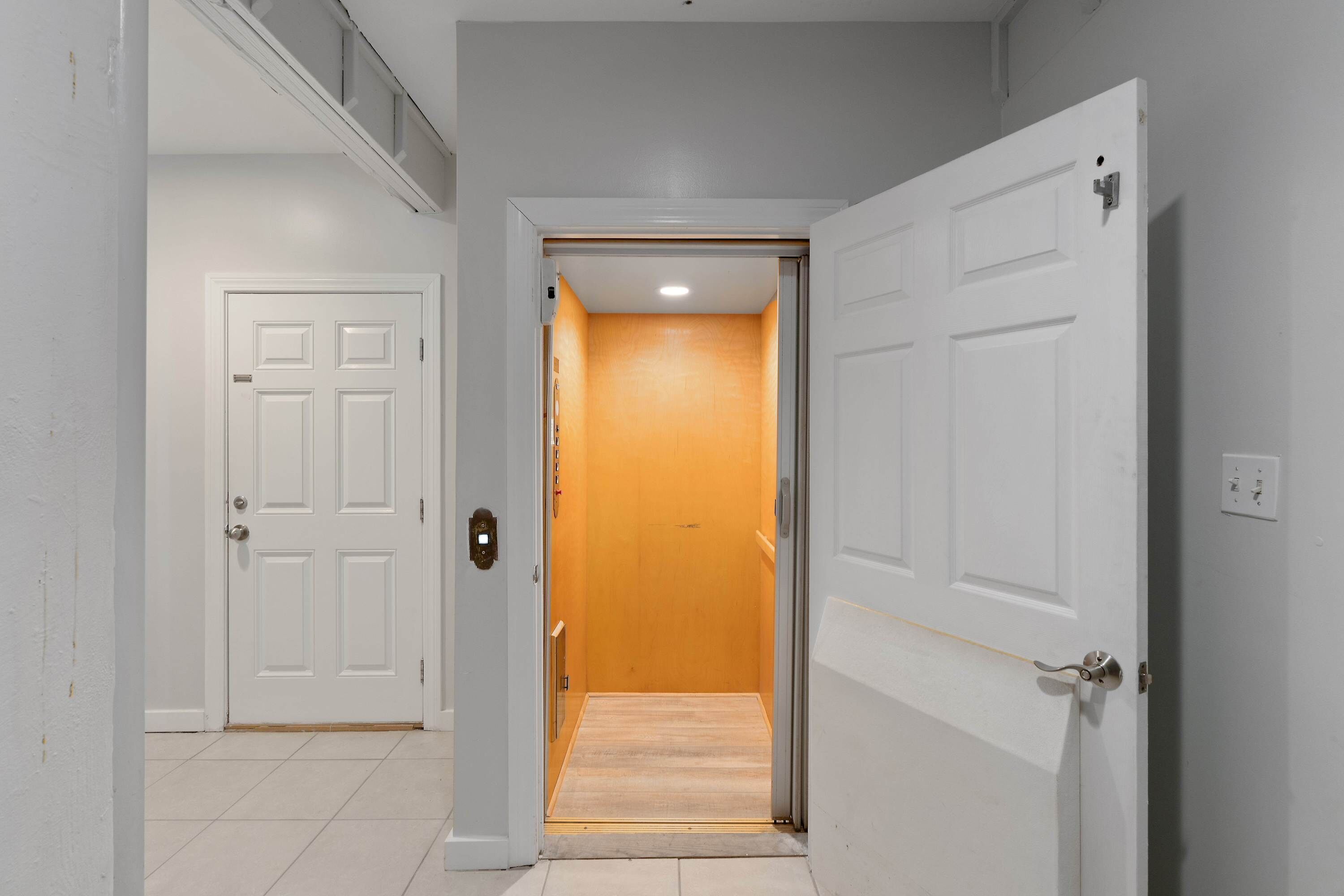 A hallway with a white door leading to a room with a wooden floor and a light fixture on the ceiling.