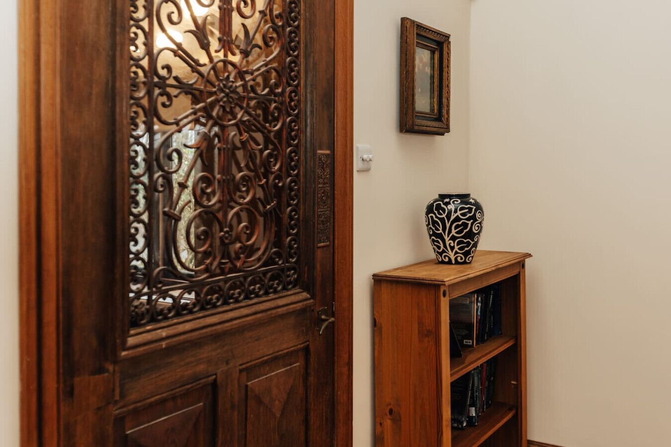 Une porte en bois ornée de motifs métalliques complexes, une petite armoire en bois surmontée d’un vase décoratif et un tableau encadré accroché au mur.