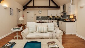 A cozy living room with a sofa, a small table with books, and a kitchen in the background. - Garden Lodge - pet friendly (Aston Tirrold)