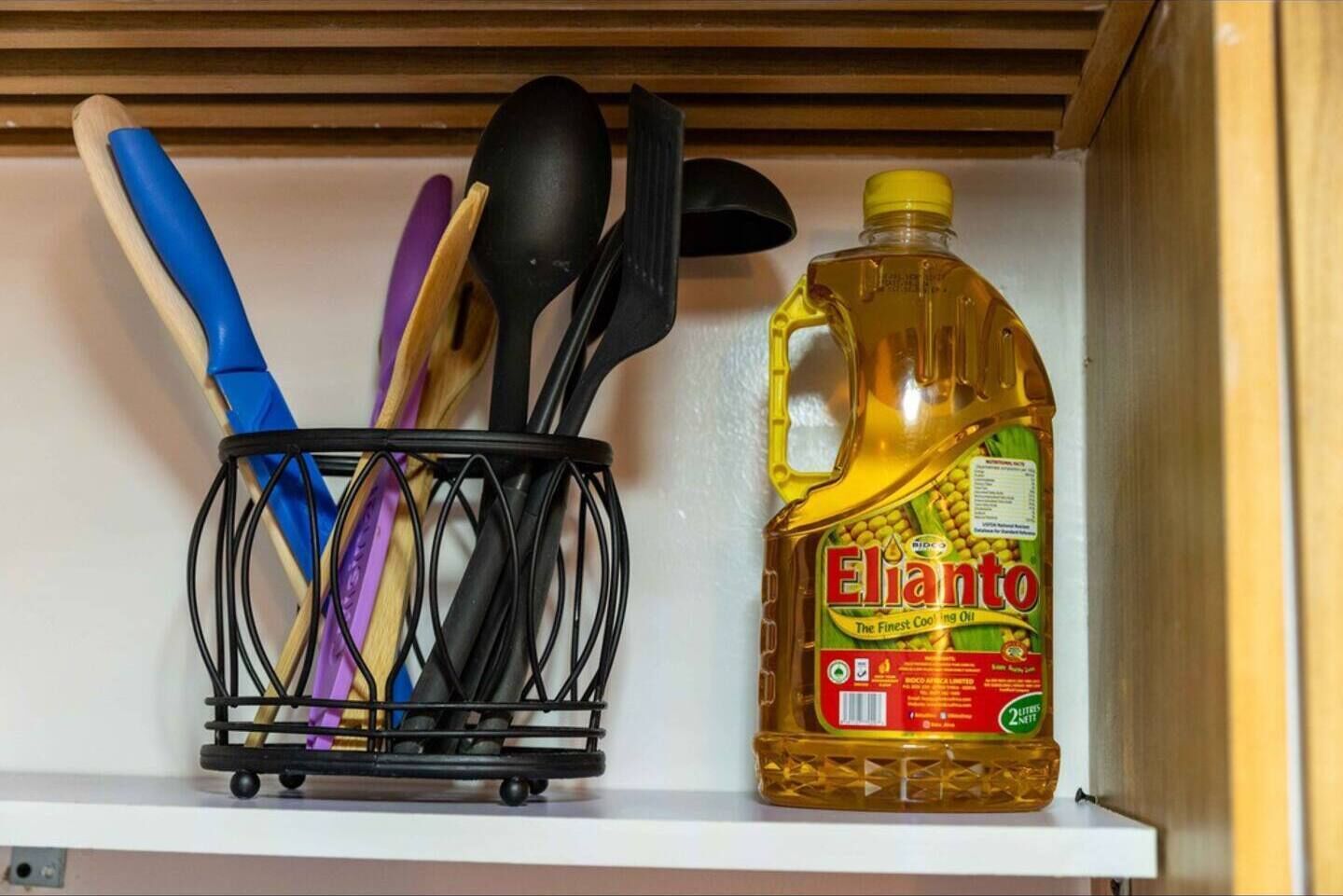 A kitchen shelf with a utensil holder containing various kitchen tools and a bottle of Elijanto cooking oil.