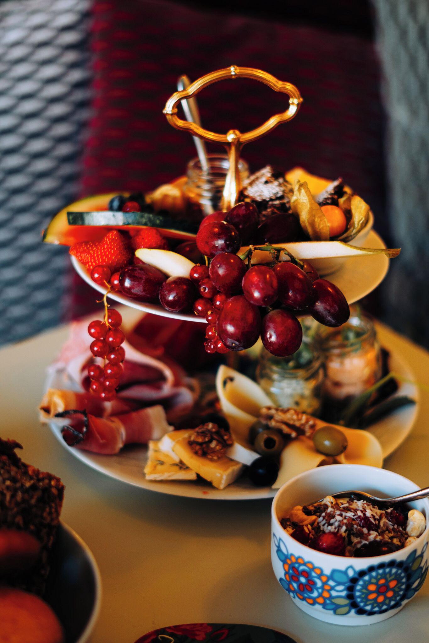 Un plato de frutas y quesos en tres niveles con un soporte metálico decorativo, un tazón de cereales y un cuenco estampado.