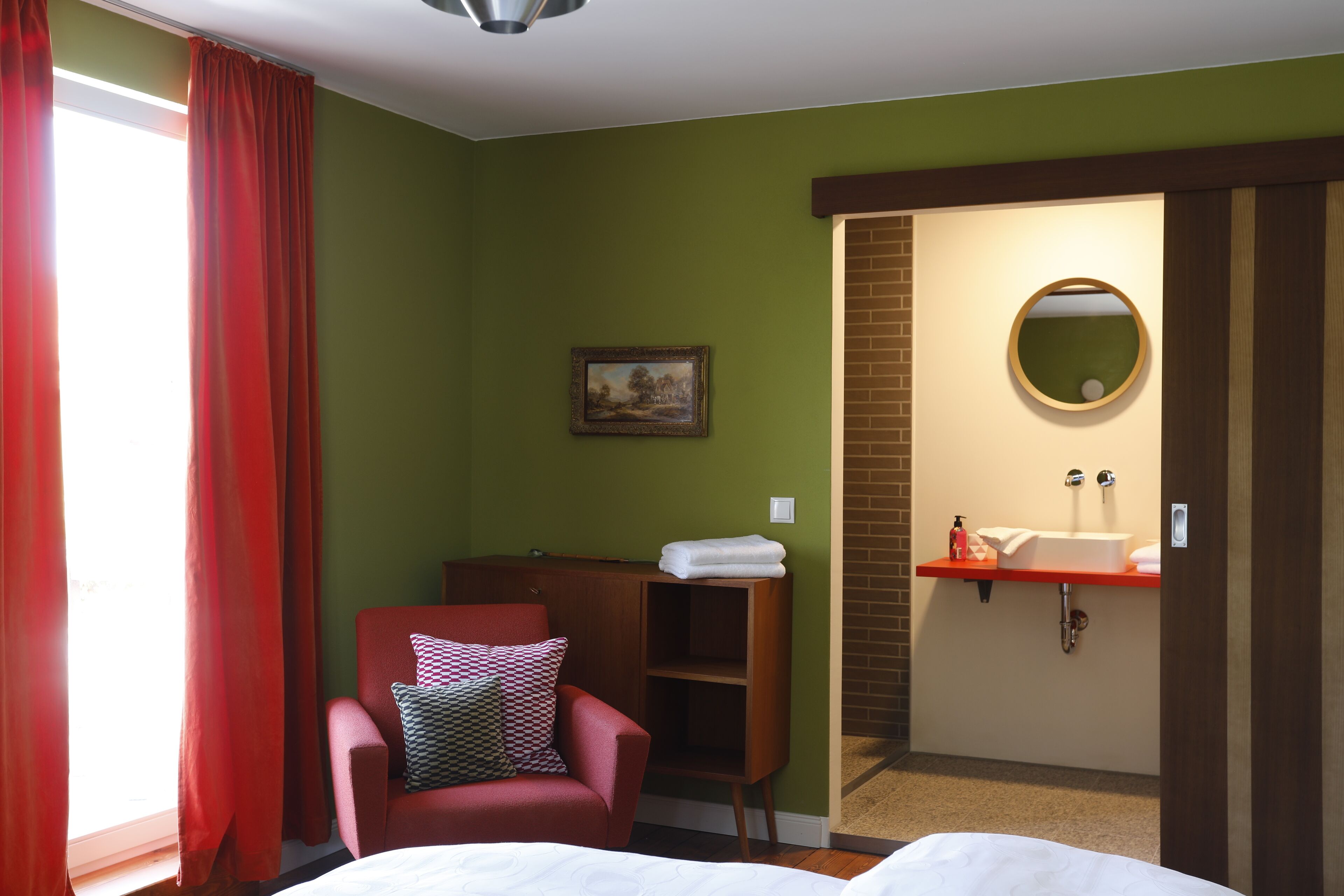 Ein Hotelzimmer mit Bett, Stuhl, kleinen Ablageflächen, einem Gemälde sowie einem Badezimmer mit Waschbecken und Spiegel.