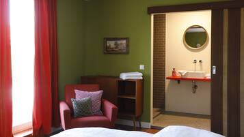 A hotel room with a bed, a chair, a small shelf, a painting, a bathroom with a sink and mirror.