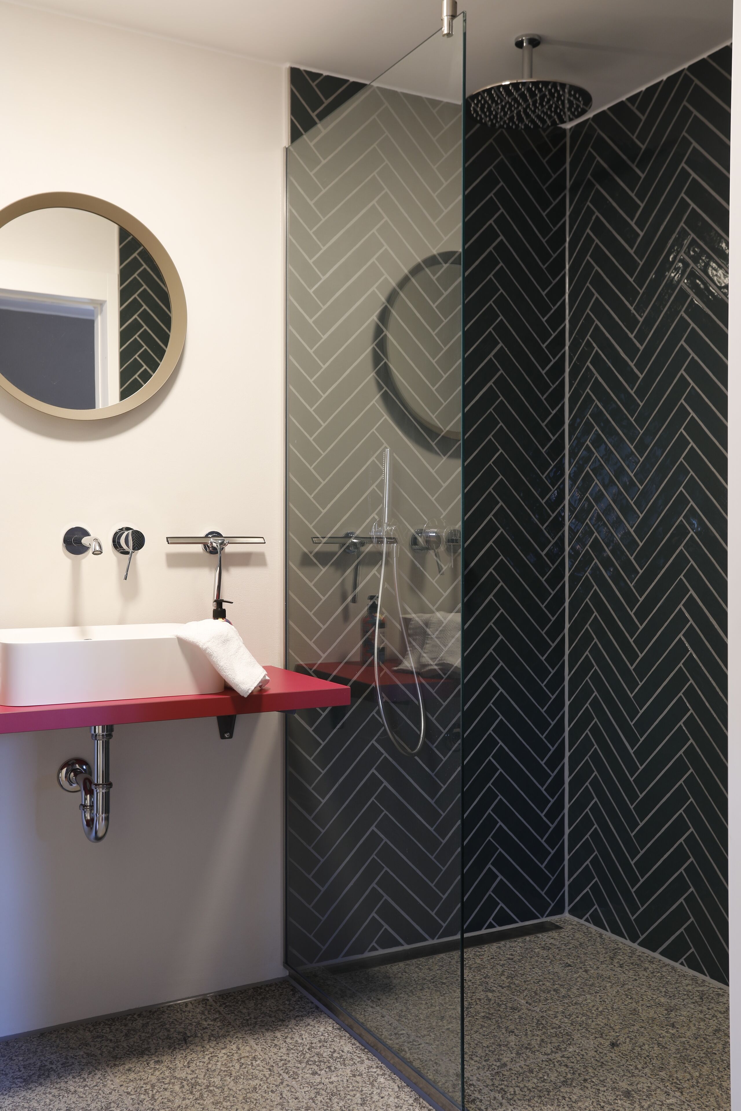 Ein modernes Badezimmer mit einer gläsernen Duschkabine, einem weißen Waschbecken mit roter Arbeitsplatte und einem Spiegel mit rundem Rahmen.