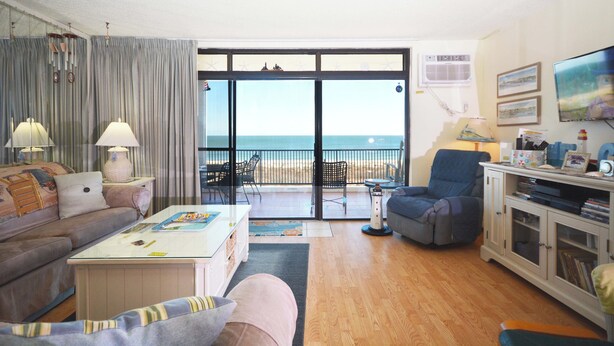 A living room with a sofa, coffee table, and armchairs, a television, and a view of the beach through large glass doors. - Exquisite, stylish 2 bedroom oceanfront condo great for families with free WiFi, an outdoor pool, and fabulous ocean view located uptown just steps to the beach! (Ocean City)