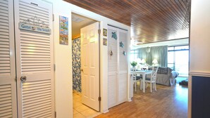 A hallway with white louvered doors, a dining area with a table and chairs, and a view of the beach through the open door. - Exquisite, stylish 2 bedroom oceanfront condo great for families with free WiFi, an outdoor pool, and fabulous ocean view located uptown just steps to the beach! (Ocean City)
